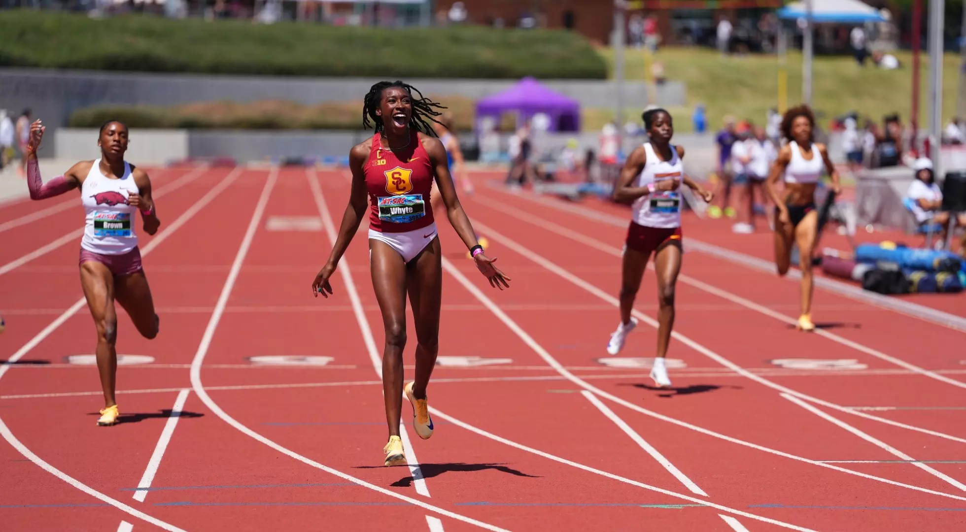 Female sprinter in USC colors leading a college track race on a red outdoor track with other athletes and stadium activity in the background
